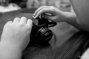Nella foto troviamo il dettaglio delle manine di una bambina fotografa che sta toccando e scoprendo per la prima volta una macchina fotografica analogica. La macchina fotografica, è impugnata con la sua mano sinistra, mentre la mano destra si sofferma per osservarla con attenzione. Lo scatto è in bianco e nero.